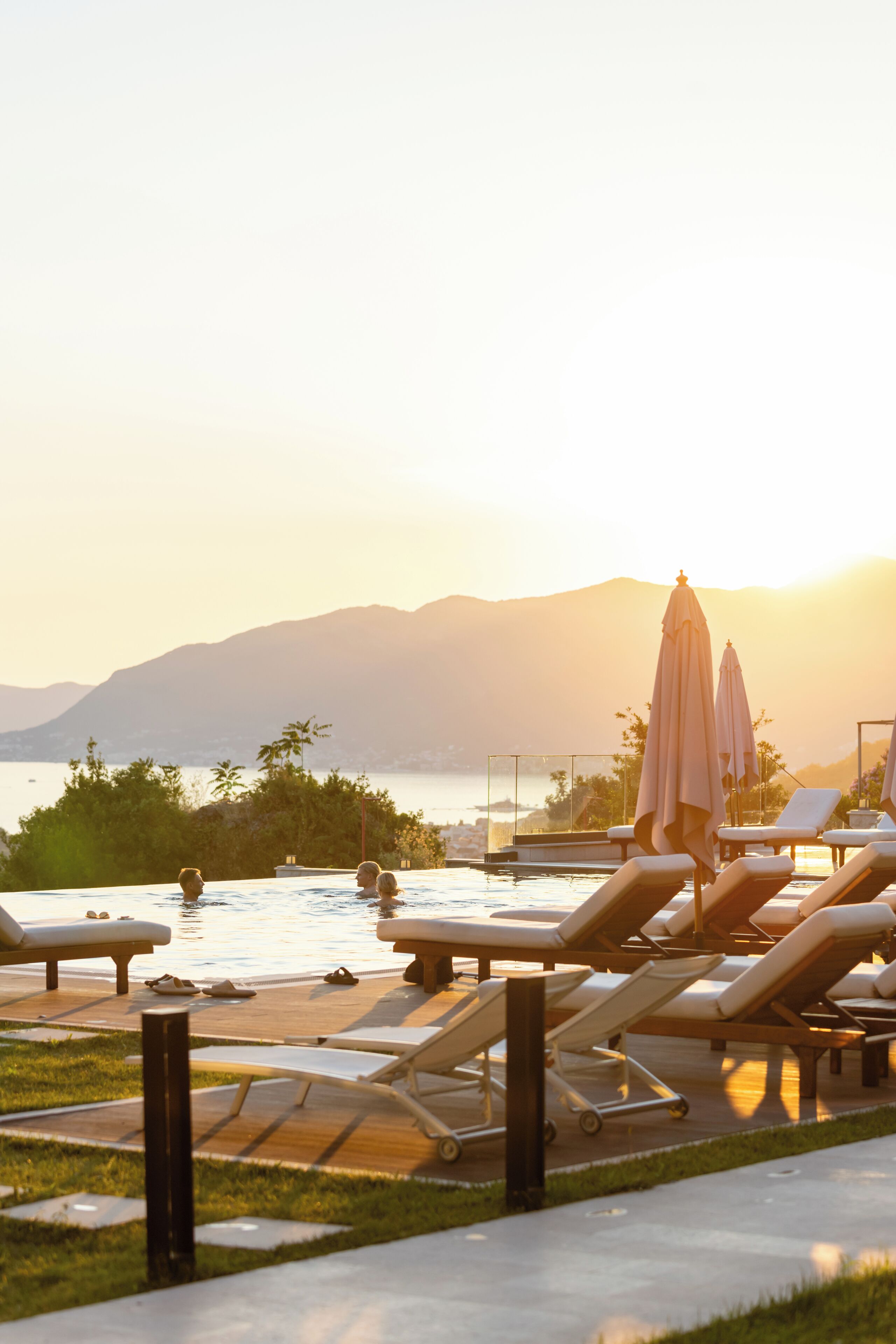 Uma área à beira da piscina com espreguiçadeiras e guarda-sóis, com vista para um lago e montanhas.