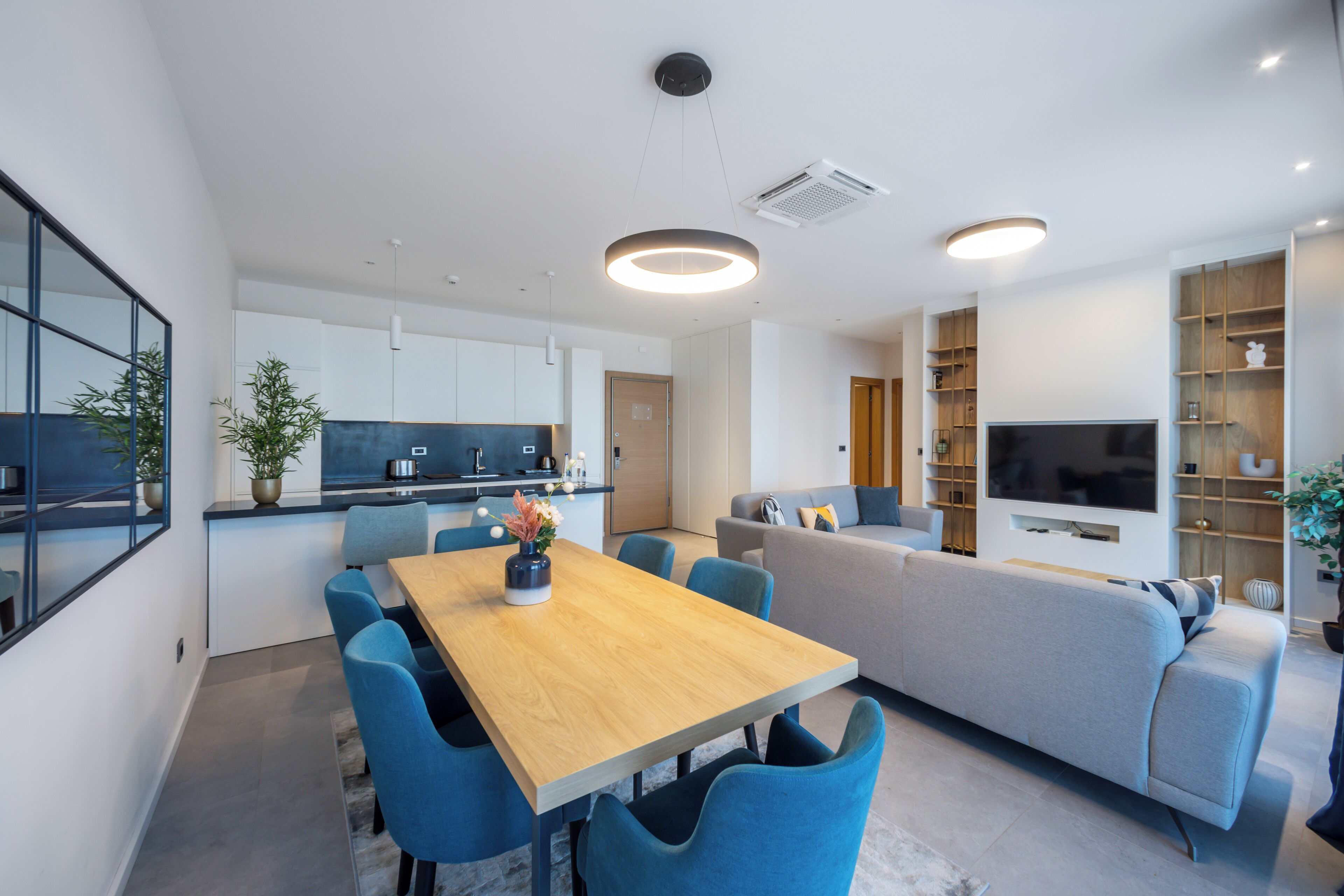 Una sala de estar moderna con una mesa de comedor de madera, sillas azules, un sofá gris y un televisor de pantalla plana.