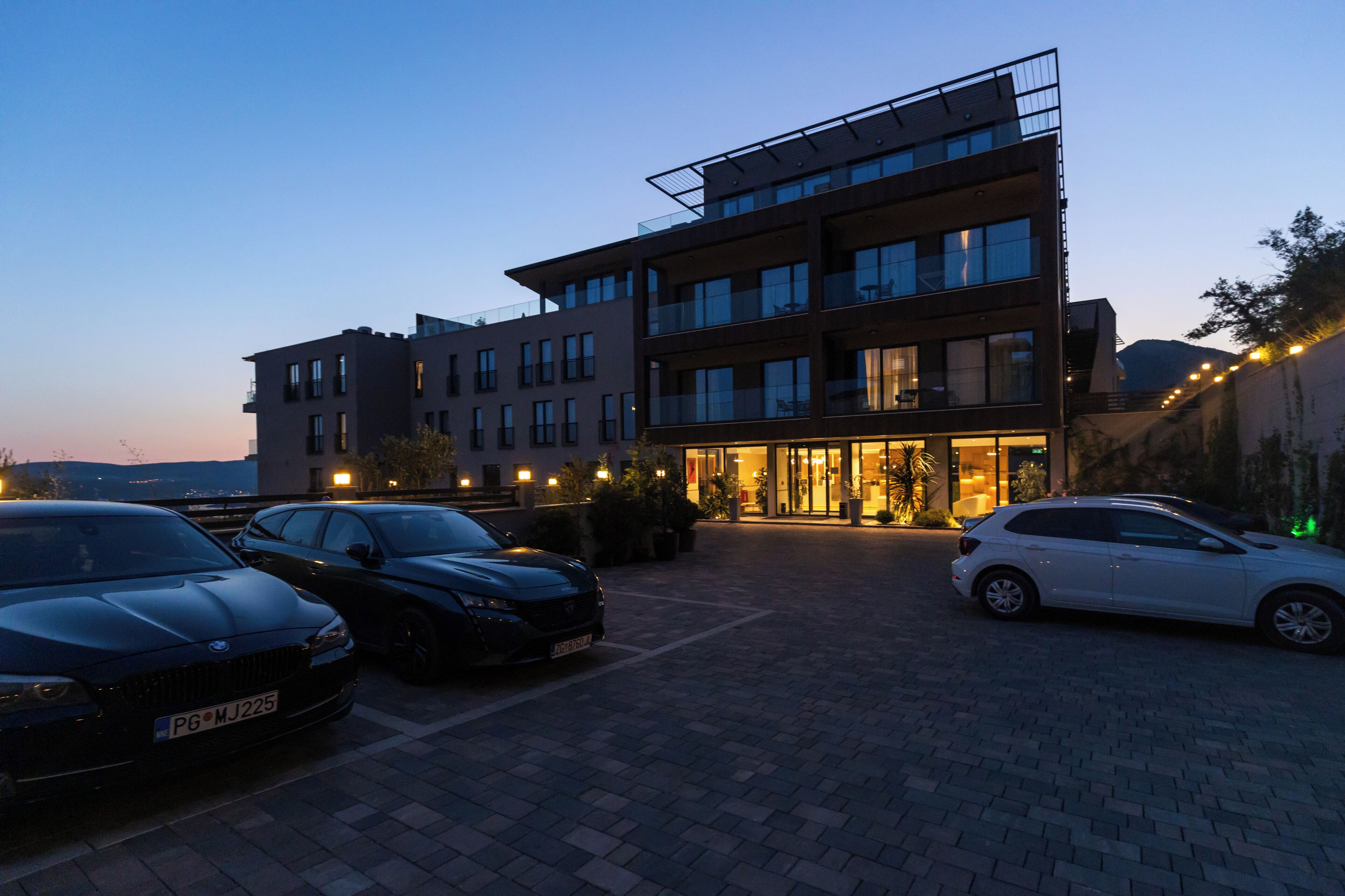 Un bâtiment moderne à plusieurs étages, un parking avec plusieurs voitures et une entrée bien éclairée.