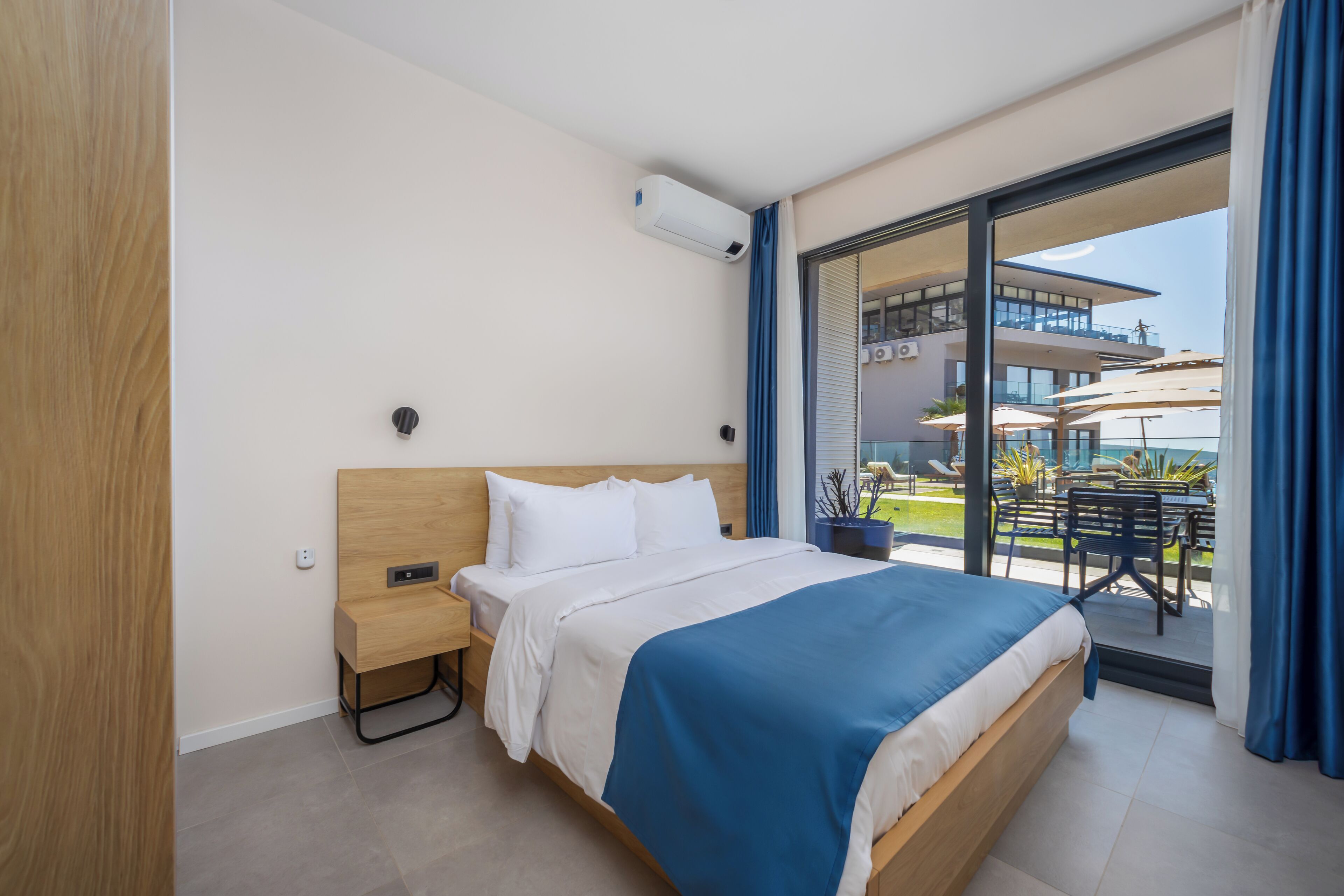 A hotel room with a large bed, a nightstand, a window with a view of the outdoor area, and a ceiling-mounted air conditioning unit.