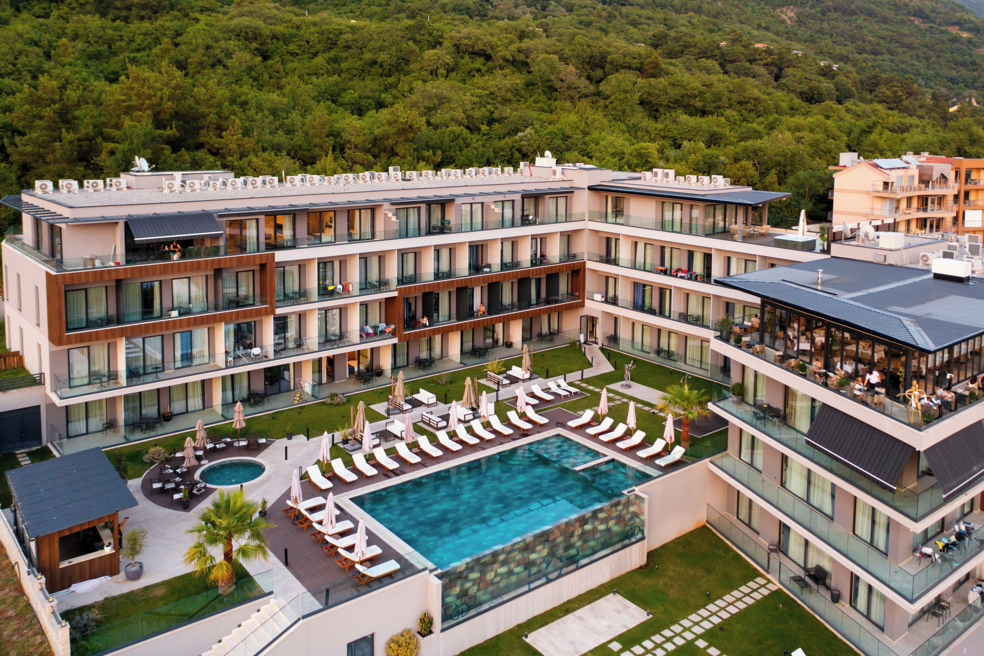 A modern hotel with a rooftop pool, multiple balconies, and a lush green landscape in the background.