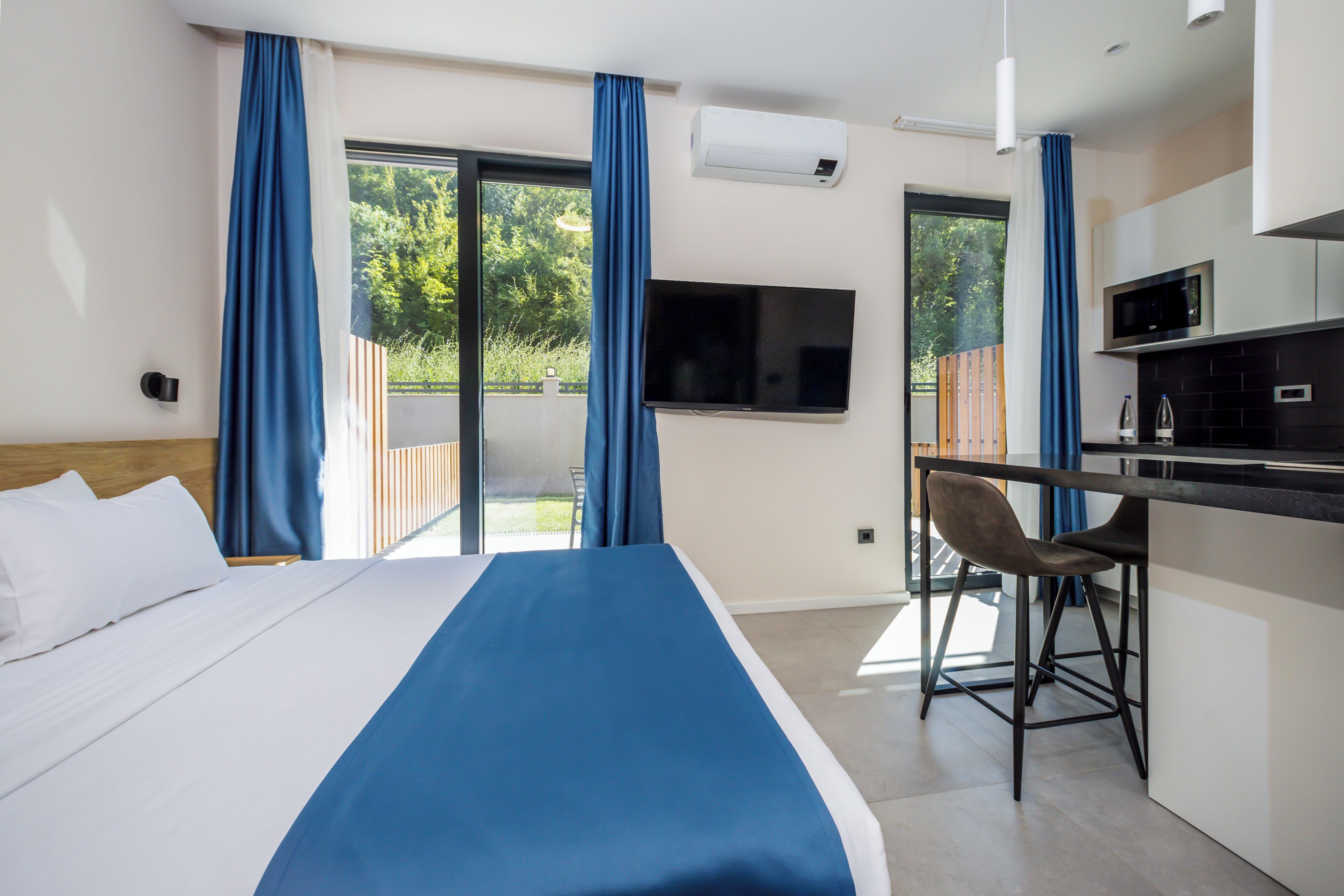 A hotel room with a bed, a desk, a chair, a TV, and a sliding glass door to the outside.