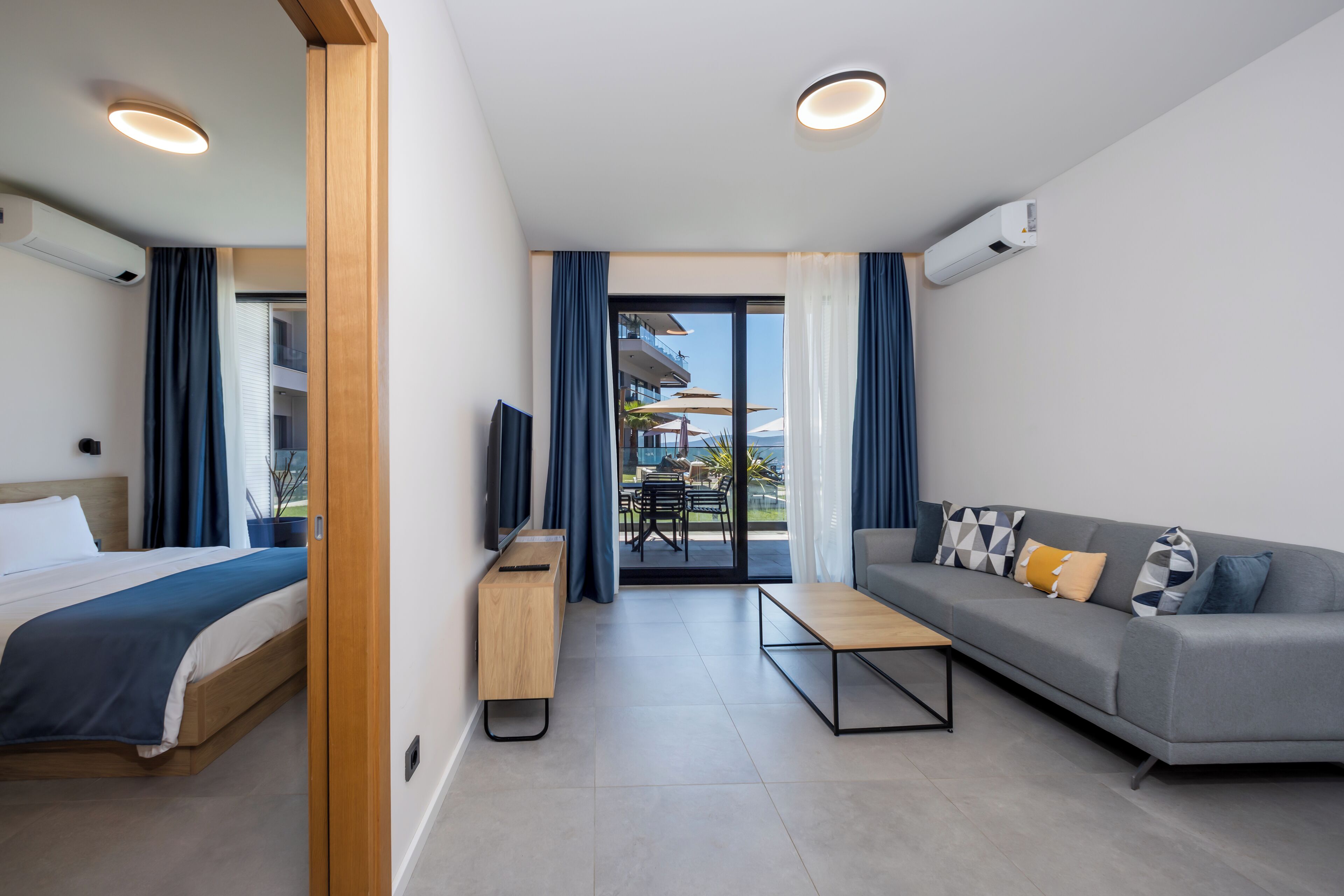 A modern hotel room with a bed, sofa, TV, and a balcony with outdoor seating.