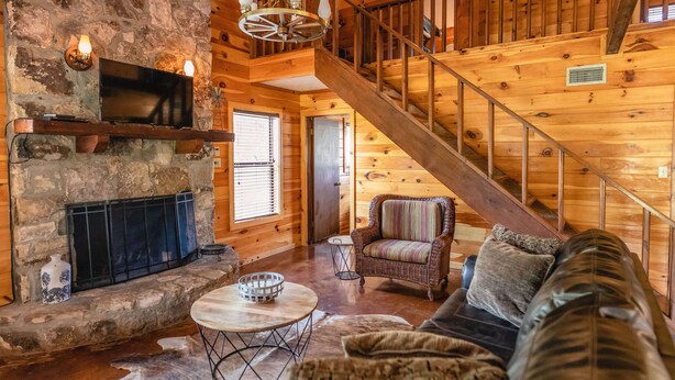 A log cabin interior with a stone fireplace, a television mounted above it, a wicker chair, a leather sofa, and a wooden staircase. - Quaint home nestled in the heart of Concan Texas! (Concan)