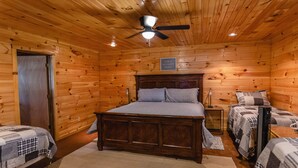 A wooden cabin bedroom with a large bed, two smaller beds, a ceiling fan, and a sign that says "THIS IS MY HAPPY PLACE". - Quaint home nestled in the heart of Concan Texas! (Concan)