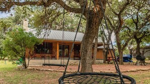 A treehouse with a swing hanging from a rope, surrounded by trees and a building in the background. - Quaint home nestled in the heart of Concan Texas! (Concan)