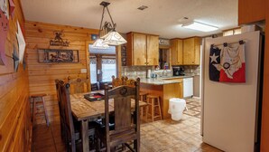 A wooden-paneled kitchen with a dining table, chairs, a hanging light fixture, wooden cabinets, a refrigerator, and a tiled floor.