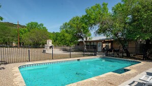 A rectangular swimming pool with a pebble-lined edge, surrounded by a fence and trees.