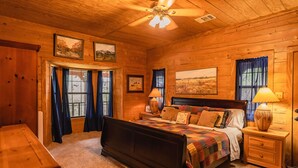 A wooden cabin bedroom with a bed, two bedside tables with lamps, a ceiling fan, and framed pictures on the walls. - Great Vacation Home with wrap around porch (Concan)