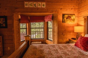 A cozy cabin room with a bed, a window with a view of trees, a wooden wall with a picture, and a nightstand with a lamp.