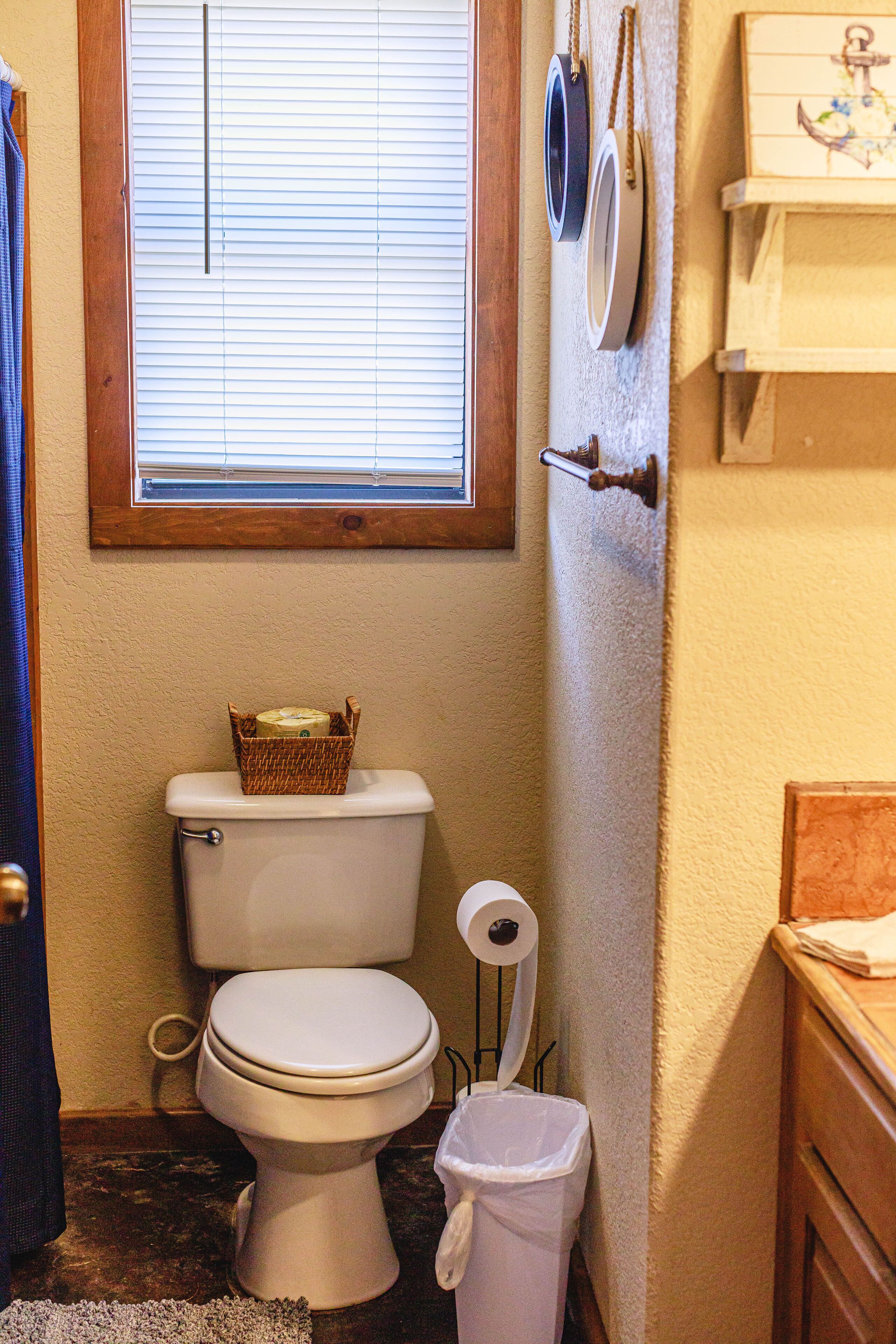 A small bathroom with a toilet, a trash can, a window with blinds, and a wooden shelf with a decorative item.