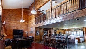 A spacious living area with a high ceiling, wooden walls, a dining area with multiple chairs, a kitchen with modern appliances, and a balcony with a railing. - Large Lodge located in Mountain Valley (Concan)