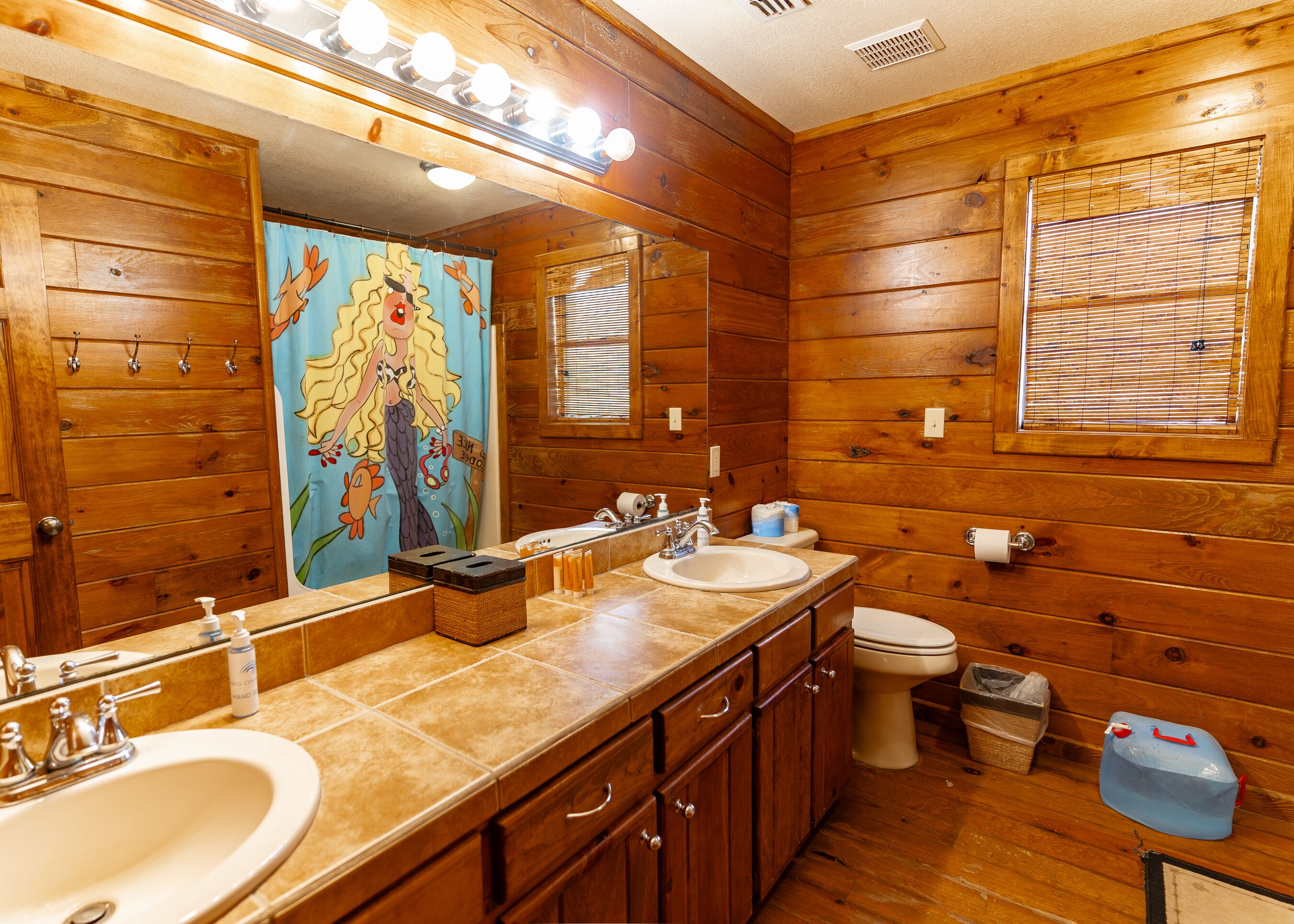 A wooden cabin bathroom with a large mirror, two sinks, a toilet, and a window with blinds.