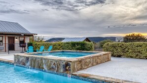 Une piscine entourée de pierres, un espace de détente couvert avec des chaises, et une vue sur des collines en arrière-plan.