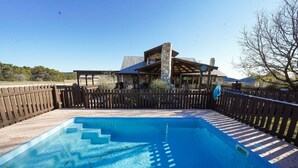 Une piscine rectangulaire avec des marches permettant d’y accéder, entourée d’une clôture en bois et bordée, au fond, d’un bâtiment en pierre.