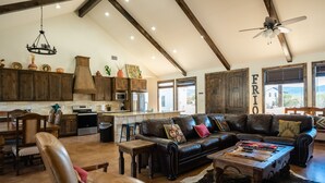 A spacious living room with a leather sofa, wooden coffee table, and a cowhide rug. A kitchen with wooden cabinets and a large window is visible in the background.