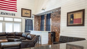 A modern living room with a leather sectional sofa, a kitchen area with a brick wall, and a large American flag. - Farmhouse - located on the 18th Hole (Concan)