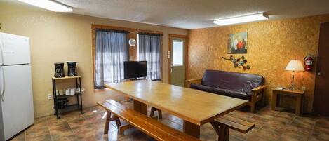 A compact living space with a kitchenette, a wooden dining table with benches, a sofa, a television, and a small side table with a lamp.