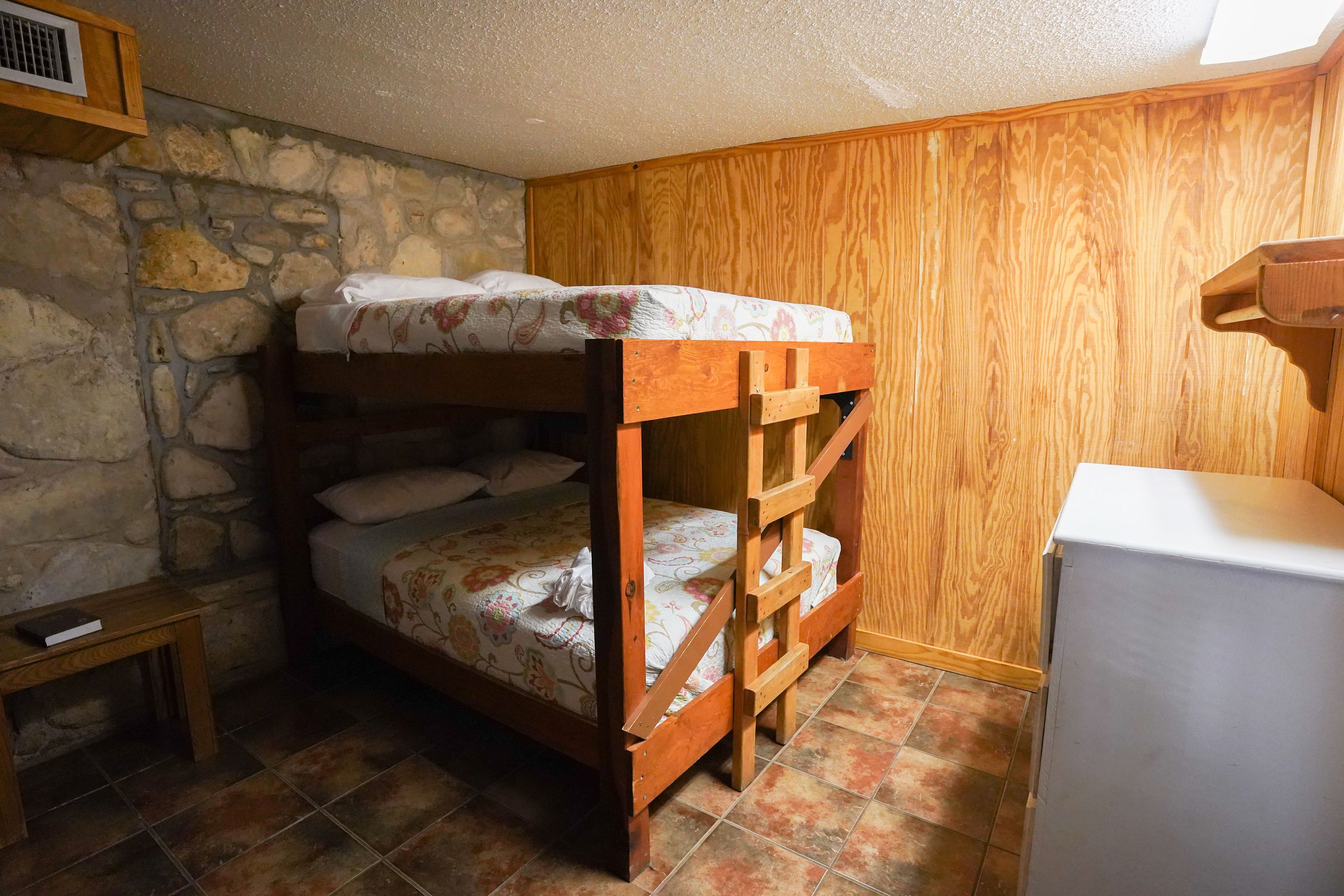 A bunk bed with a wooden frame and floral bedding, a small wooden table with a book, and a stone wall.
