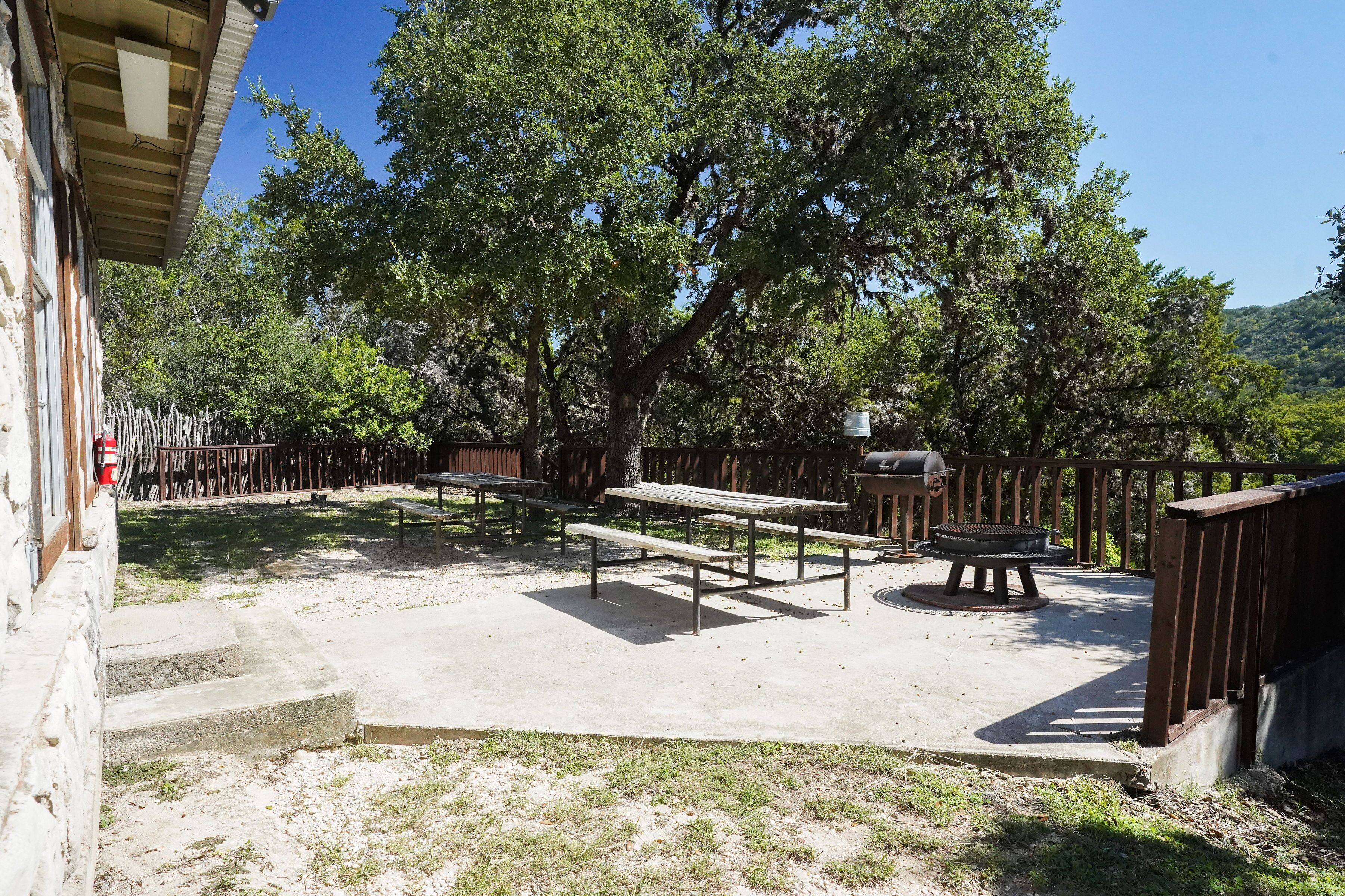 An outdoor patio area with picnic tables and a barbecue grill, surrounded by trees and a stone wall.