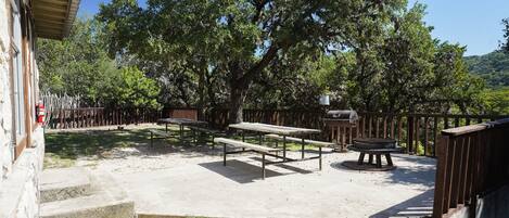 An outdoor patio area with picnic tables and a barbecue grill, surrounded by trees and a stone wall.