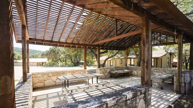 An outdoor covered patio area with stone walls and wooden beams, featuring several benches and tables. - Quaint cabin located near the Frio River (Concan)