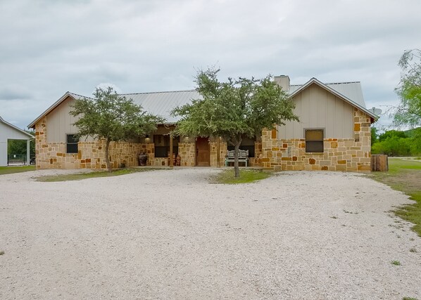 A two-story stone building with a gravel driveway and a large tree in front. - Nestled in Tierra Linda (Concan)
