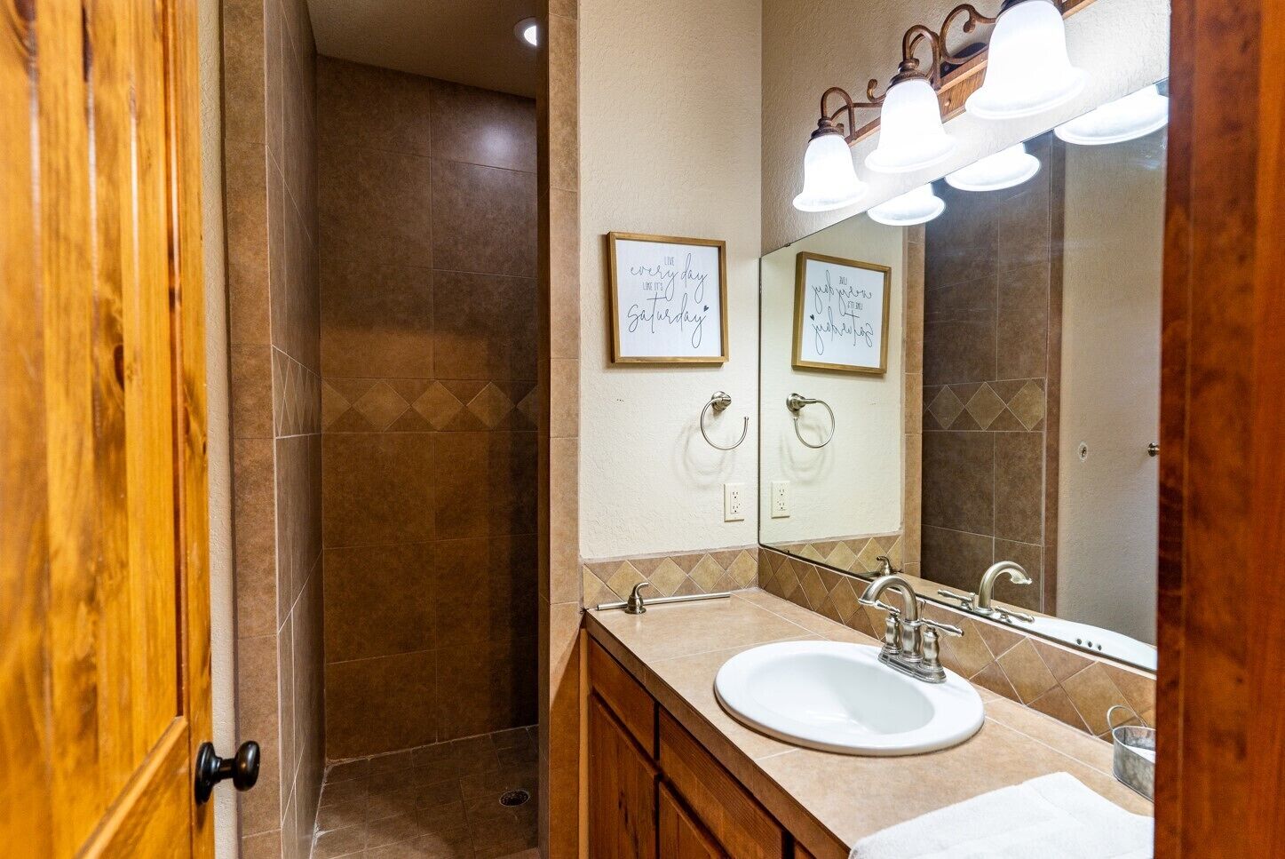 A bathroom with a wooden door, a white sink, a mirror with light fixtures, and a shower area.