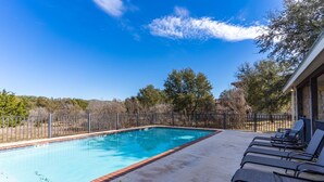 A rectangular swimming pool with a tiled edge, surrounded by a concrete deck and a metal fence. There are lounge chairs along the deck's edge.