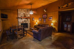 A cozy living room with a stone fireplace, leather sofa, wooden furniture, and wall-mounted decorations. - River Front cabin with access to shared pool! (Concan)