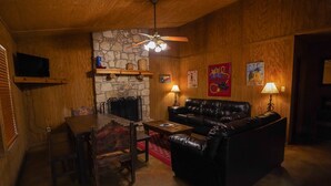 A cozy living room with a stone wall, wooden ceiling, a black leather sofa, a wooden dining table with chairs, and a ceiling fan. - River Front cabin with access to shared pool! (Concan)