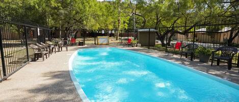A rectangular swimming pool with a tiled edge, surrounded by a black metal fence. There are several lounge chairs and a red chair near the pool.