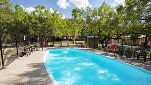 Ein rechteckiger Swimmingpool mit einer gefliesten Kante, umgeben von einem schwarzen Metallzaun. Es befinden sich mehrere Liegestühle und ein roter Sessel in der Nähe des Pools.