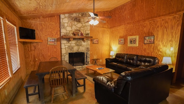 A cozy living room with a stone fireplace, wooden walls, a leather sofa, a dining table with chairs, and a ceiling fan. - Walking distance to the Frio River, access to shared pool (Concan)