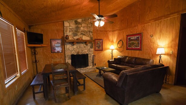 A cozy living room with wooden walls, a stone fireplace, a leather sofa, a wooden table with chairs, and a ceiling fan. - Walking distance to the Frio River, access to shared pool (Concan)