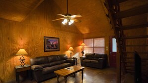Una sala de estar acogedora con paredes de madera y techo inclinado, que incluye dos sofás de cuero, una mesa de centro de madera y un ventilador de techo.