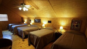 Quarto com quatro camas dispostas em linha, paredes de madeira, ventilador de teto e um quadro na parede.