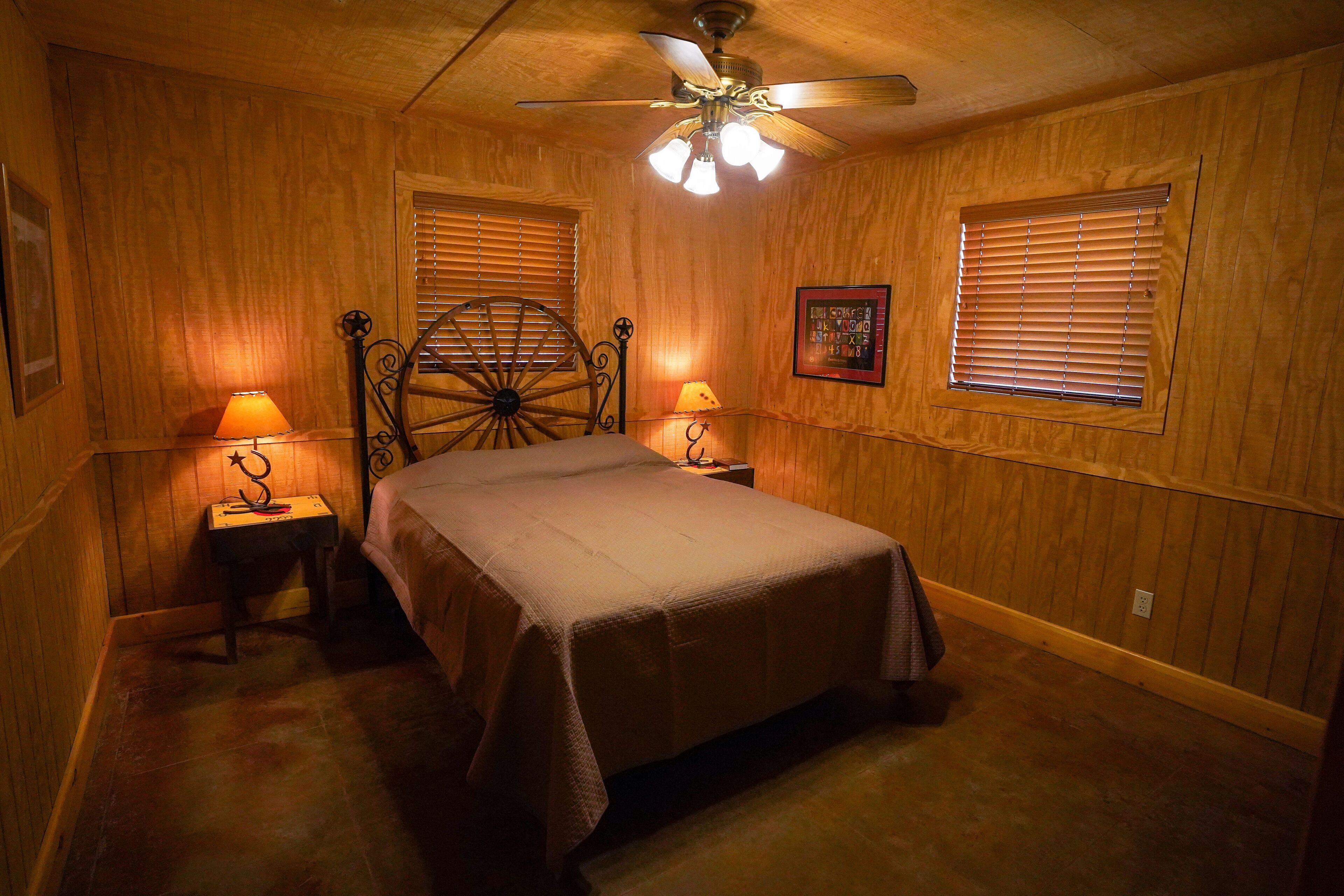 Un dormitorio con cama, dos mesitas de noche con lámparas, un ventilador de techo, paredes de madera y una ventana con persianas.