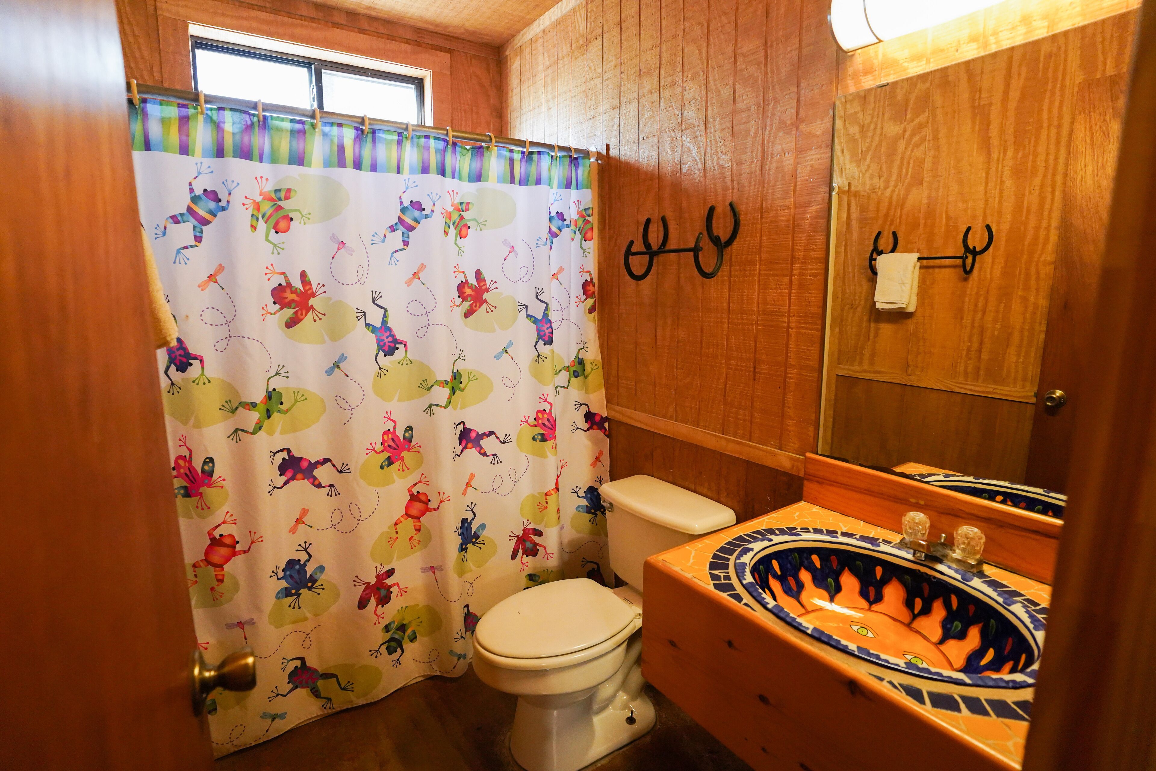 A bathroom with a white toilet, a colorful shower curtain with frog patterns, a wooden vanity with an orange and blue sink, and a mirror with a towel hanging on a metal hook.