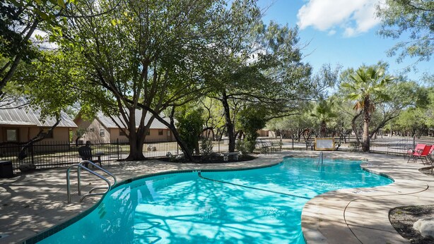 A swimming pool with a curved edge, surrounded by trees and a fence. - Riverfront cabin with access to shared pool! (Concan)