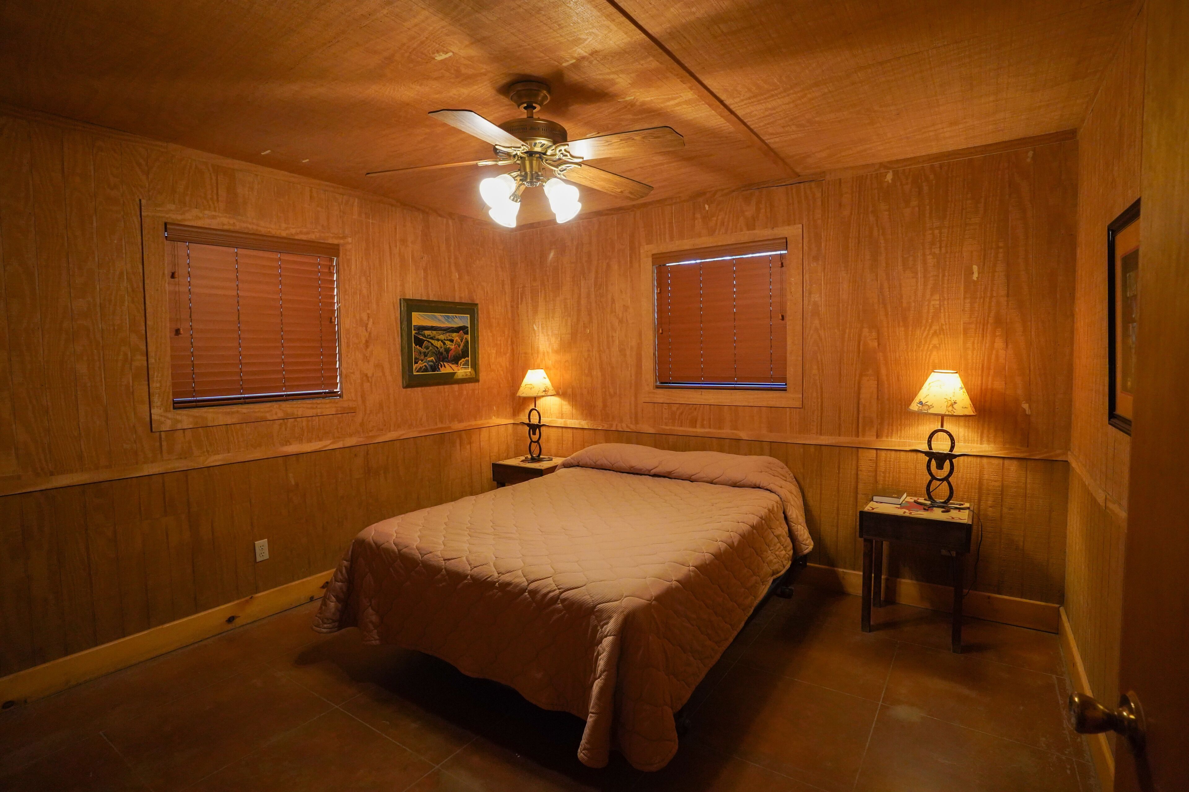 Une chambre avec des murs en bois, un ventilateur de plafond, deux lampes de chevet et un lit recouvert d’une couverture marron.