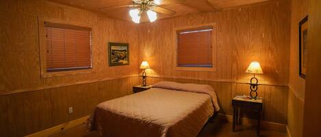 Une chambre avec des murs en bois, un ventilateur de plafond, deux lampes de chevet et un lit recouvert d’une couverture marron.