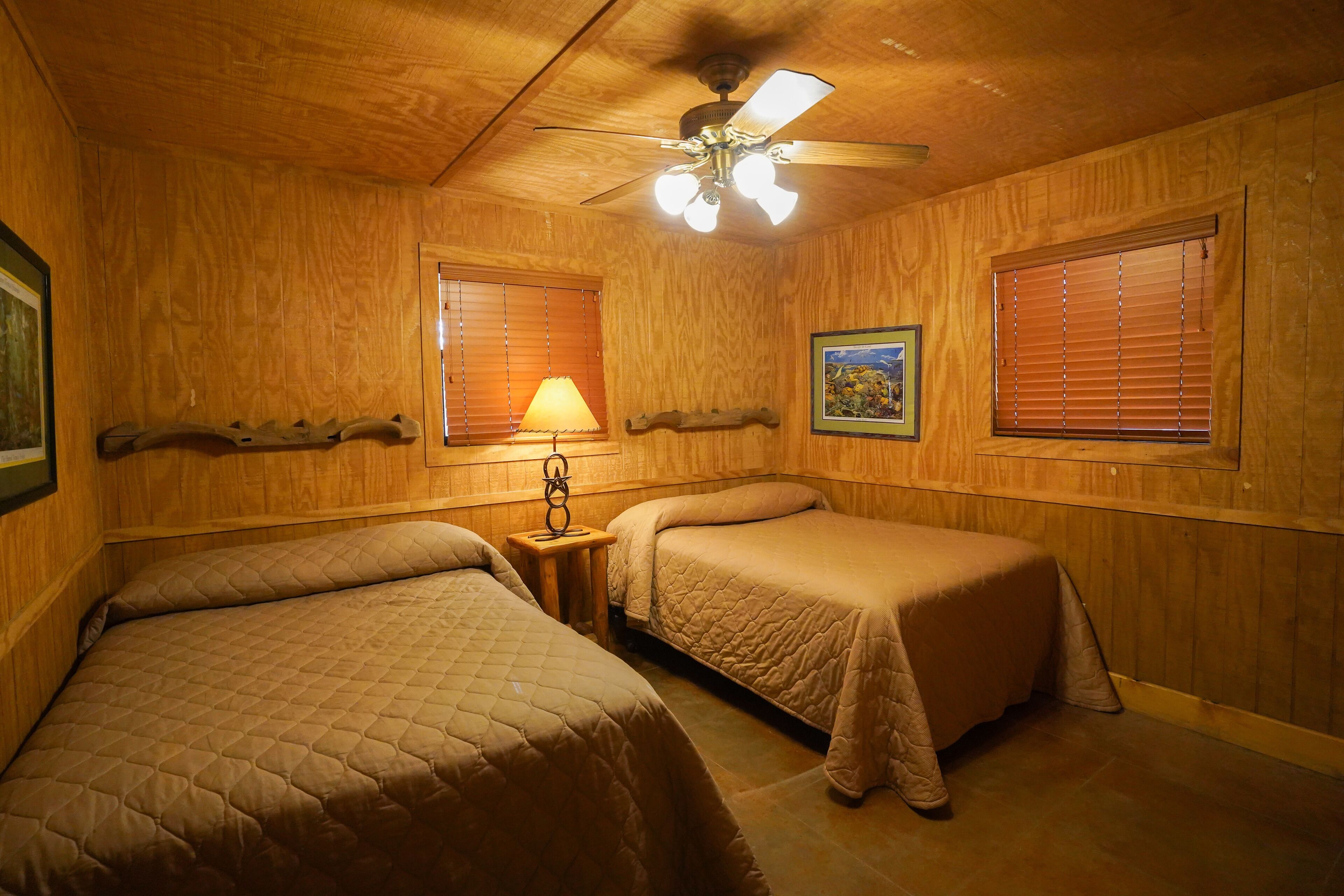 Ein Zimmer mit Holzwänden, zwei Betten mit Bettdecken, einem Deckenventilator, einer Lampe und zwei Fenstern mit Jalousien.