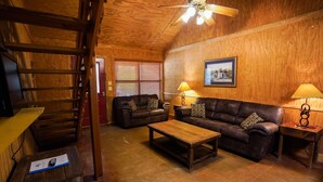 A cozy living room with wooden walls, a ceiling fan, a wooden coffee table, two leather sofas, and two lamps. - Riverfront cabin with access to shared pool! (Concan)