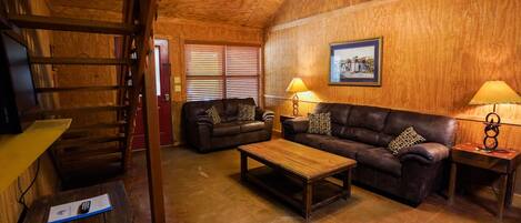 A cozy living room with wooden walls, a ceiling fan, a wooden coffee table, two leather sofas, and two lamps.