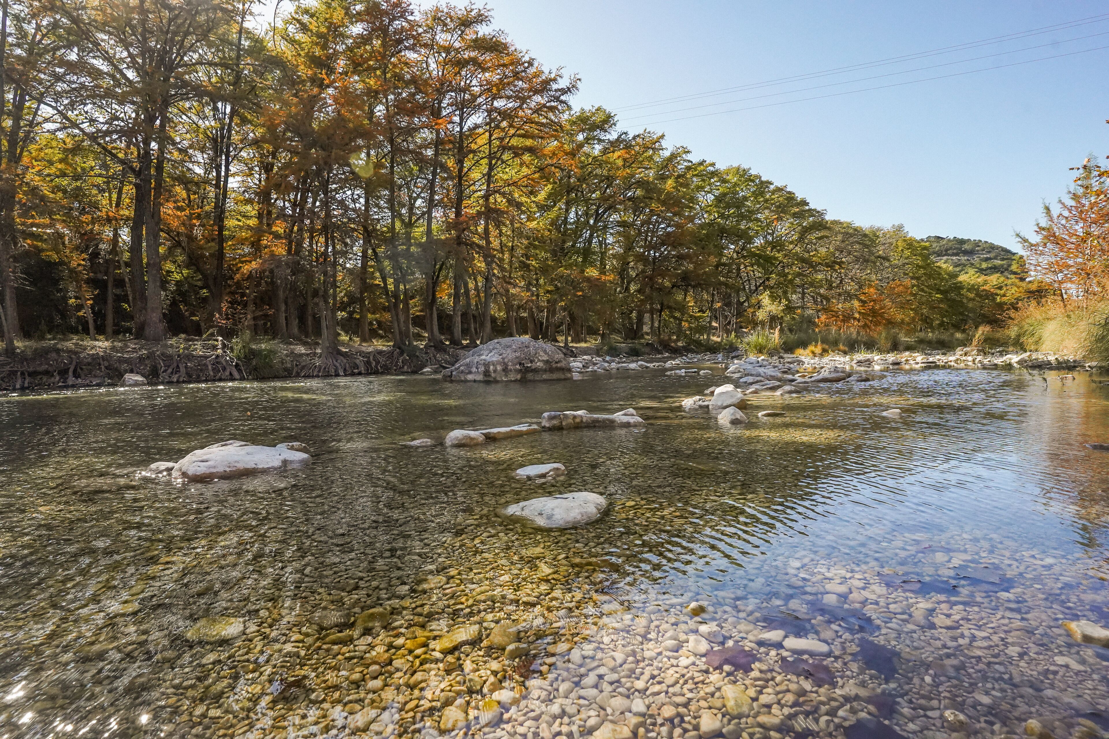 A river with clear water and rocks, surrounded by trees with autumn foliage.