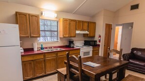 A compact kitchen with wooden cabinets, a refrigerator, a stove, and a microwave. There's a dining table with chairs and a leather couch in the adjacent living area. - Frio Country Resort - Within walking distance to the Frio River! (Concan)