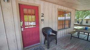 A wooden porch with a red door, a bench, and a table. - Frio Country Resort - Within walking distance to the Frio River! (Concan)