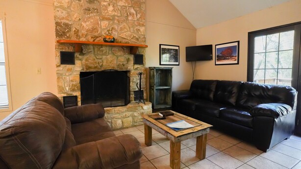 A living room with a stone fireplace, a black leather sofa, a wooden coffee table, and a television mounted on the wall. - Frio Country Resort - Within walking distance to the Frio River! (Concan)
