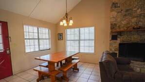 A dining area with a wooden table and benches, large windows with blinds, a stone fireplace, and a red door. - Frio Country Resort - Within walking distance to the Frio River! (Concan)
